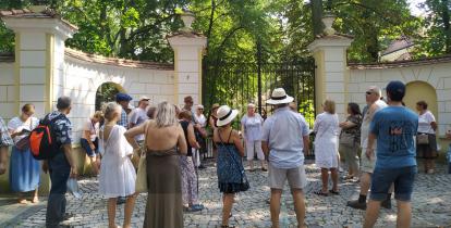 Grupa starszych i młodszych osób stoi zwrócona do jednej kobiety. Za nimi wysokiemurowane ogrodzenie z bramą pośrodku. Za bramą rosna liściaste drzewa.  Jest słoneczna pogoda. 