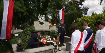 Dwóch strażników miejskich pełni wartę przy nagrobku na cmentarzu. Przed nageobkiem młodzi ludziewbiało-czarnych strojach. 