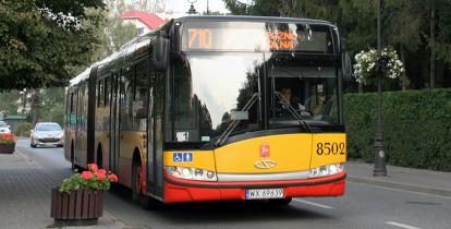 Ulica. Na niej stoi żółto-czerwony autobus komunikacji miejskiej. Z przodu na górze jest napis 710 Piaseczno.  Obok chodnik dla pieszych i donice z kwiatami.