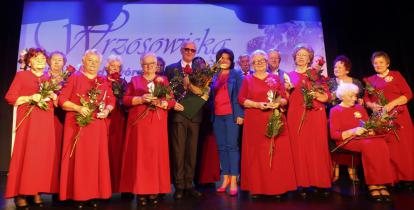 Grupa starszych kobiet w czerwonych sukniach stoi na scenie, a wśród nich mężczyzna w garniturze i kobieta w niebieskiej marynarce i spodniach.