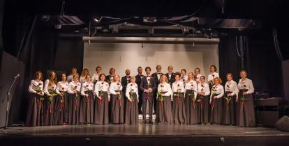 Zdjęcie grupowe Konstancińskiego Chóru Kameralnego. Osiemnaście osób stoi w dwóch rzędach. Ubrani są na czarno-biało. Członkowie chóru stoją na scenie.