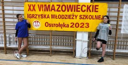 Dwie uczennice spozuja na tle baneru informującym o 26. Mazowieckich Igrzyskach Młodzieży Szkolnej.