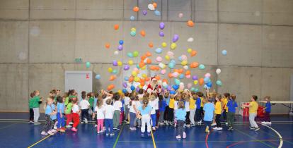 Przedszkolaki stoją na hali sportowej zebrane do okoła Mikołaja. Nad nimi lataja balony. 