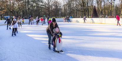 Park zdrojowy, ludzie w strojach zimowych jeżdżą na lodowisku, w tle budynek amfiteatru i drzewa.