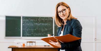  Kobieta – nauczycielka stoi w klasie. W rękach trzyma duży zeszyt. W tle widać tablicę oraz biurko.