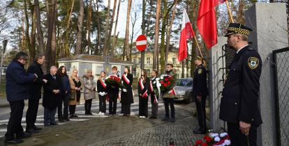 Grupa młodych dziewczyn i chłopców z biało-czerwonymi wieńcami i szarfami oraz trzech starszych mężczyzn i trzy kobiety stoją naprzeciwko tablicy zawieszonej na ogrodzeniu. Przy ogrodzeniu dwóch strażników miejskich w galowych mundurach oraz biało-czerwone flagi.