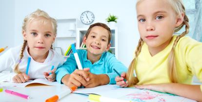 Dwie dziewczynki z blond warkoczykami, a w środku chłopiec. Siedzą za stołem w szkolnej klasie i rysują.