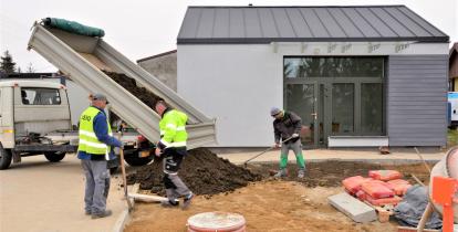 Budowa, wywrotka zrzuca ziemie, 3 robotników stoi obok. W tle widać szary budynek. 