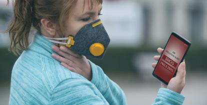 Kobieta z maseczką na twarzy patrzy w monitor telefonu, na który wyświetlony jest napis " Air pollution alert: Smog"