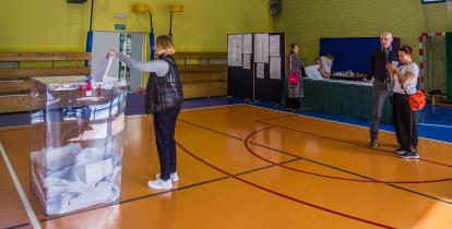 Pomieszczenie sali gimnastycznej, kobieta wrzuca kartkę do urny wyborczej 
