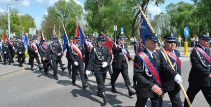 Strażacy maszerują ulicą. Za nimi zielone drzewa. W dłoniach trzymają sztandary.