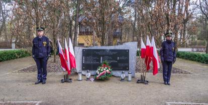 Pośrodku czarna tablica granitowa z tekstem umocowana na prostokątnym kamieniu, po lewej i prawej stronie stoją flagi narodowe, przed tablicą leżą kwiaty i wiązanki oraz palą się znicze