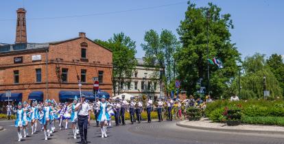 Orkiestra dęta idącą na czele parady, w tle budynek z czerwonej cegły z kominem.