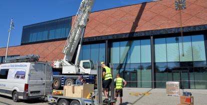 Przed budynkiem z czerwonej cegły grupa osób rozładowuje towar.