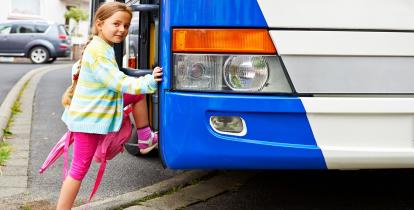 Dziewczynka ubrana w różową sukienkę i zielony sweter wsiada do biało-niebieskiego autobusu.