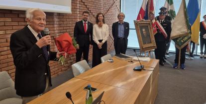 Starszy mężczyzna trzyma w rękach mikrofon i kwiaty.