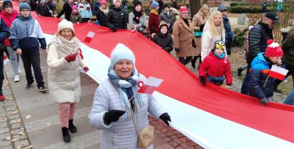 Grupa osób, w tym dzieci i osoby starsze, bierze udział w marszu z okazji Święta Niepodległości. Uczestnicy trzymają dużą, biało-czerwoną flagę Polski, a niektórzy mają przypięte kotyliony.