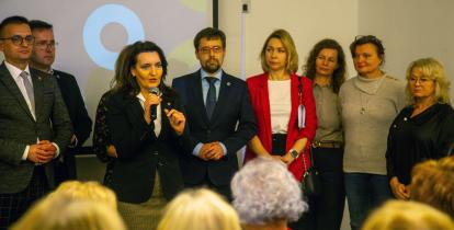 Grupa osób stoi na scenie, a kobieta w ciemnym żakiecie przemawia do mikrofonu przed publicznością.