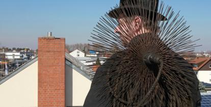 Mężczyzna, kominiarz stoi na dachu. Ubrany jest w czarną kurtkę, na głowie ma czarny kapelusz. Na plecy ma zarzucone urządzenie do czyszczenia kominów z drutami ułożonymi w okrąg. W tle domy jednorodzinne i błękitne niebo.