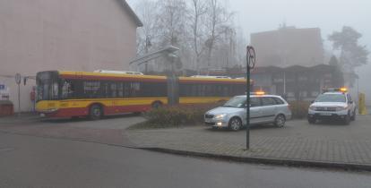 Żółto-czerwony autobus przegubowy warszawskiej komunikacji miejskiej zaparkowany na pętli autobusowej, w otoczeniu budynków i pawilonów handlowych spowitych mgłą. Obok stoją dwa pojazdy techniczne Zarządu Transportu Miejskiego z włączonymi światłami ostrzegawczymi.