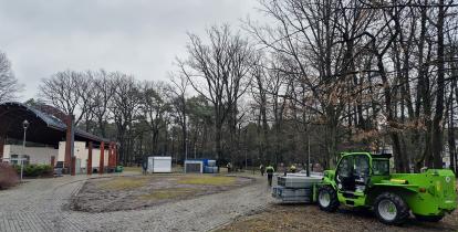 park z zadaszoną sceną, robotników w kamizelkach pracujących przy montażu lodowiska oraz zieloną ładowarkę teleskopową transportującą elementy konstrukcyjne.