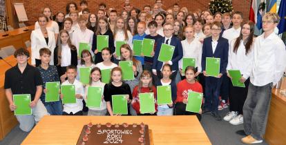 Grupa młodych sportowców trzymających zielone dyplomy pozuje do zdjęcia w sali konferencyjnej z dekoracyjną choinką i tortem z napisem Sportowiec Roku 2024 na pierwszym planie.