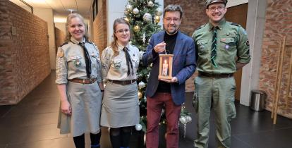 Grupa czterech osób stoi przed udekorowaną choinką, z czego trzy osoby są w mundurach harcerskich, a jedna trzyma zapaloną lampkę.