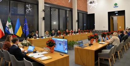 Zdjęcie przedstawia salę obrad z uczestnikami siedzącymi przy stołach, udekorowaną flagami Polski, Ukrainy, Unii Europejskiej oraz roślinami w świątecznym wystroju.