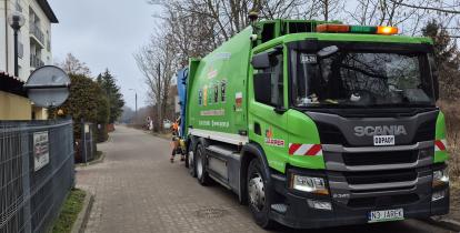 Zielona śmieciarka firmy Jarper marki Scania, stojąca na osiedlowej uliczce podczas odbioru odpadów przez pracownika w pomarańczowym stroju.