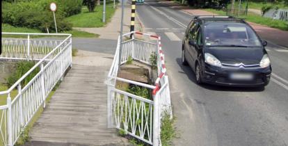 Widok na drewnianą kładkę przez rów wodny, obok samochód.
