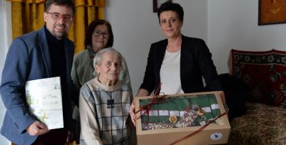Grupa osób, w tym jubilatka, otrzymuje upominki i gratulacje z okazji setnych urodzin w domowym otoczeniu.