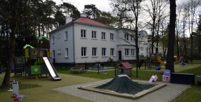 Budynek Przedszkola nr 1 „Zielony Zakątek” otoczony drzewami, z placem zabaw wyposażonym w zjeżdżalnię, piaskownicę i huśtawki.