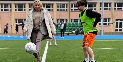 Kobieta w eleganckim stroju kopie piłkę na boisku sportowym, podczas gdy chłopiec w sportowym stroju obserwuje ją z uwagą.