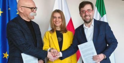 Trzech przedstawicieli podpisuje porozumienie, uśmiechając się i ściskając dłonie na tle flag Unii Europejskiej, Polski i Konstancina-Jeziorny.