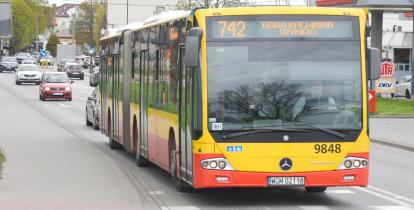 Przegubowy autobus Mercedes-Benz w żółto-czerwonych barwach warszawskiej komunikacji miejskiej, kursujący na linii 742 do Góry Kalwarii, jadący po miejskiej drodze wśród innych pojazdów.
