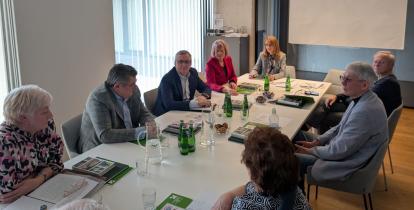 Grupa osób uczestniczy w formalnym spotkaniu przy stole konferencyjnym, omawiając dokumenty i notatki.