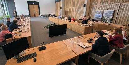 Spotkanie w nowoczesnej sali konferencyjnej, gdzie uczestnicy siedzą przy długich drewnianych stołach, wyposażonych w mikrofony i monitory, uważnie słuchając przemawiających osób.