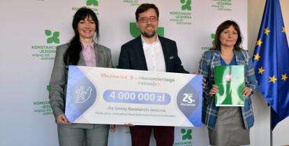 Trzy osoby pozują do zdjęcia, trzymając symboliczny czek na 4 mln zł dofinansowania dla Gminy Konstancin-Jeziorna na modernizację stacji uzdatniania wody Nowe Wierzbno; w tle logo gminy i flaga Unii Europejskiej.