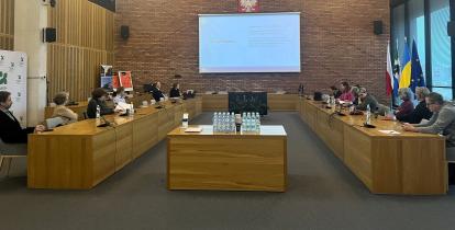 Spotkanie konsultacyjne w sali obrad, podczas którego uczestnicy omawiają strategię rozwoju gminy, a na ekranie wyświetlany jest plan spotkania.