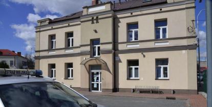 Piętrowy budynek straży miejskiej, na pierwszym planie fragment samochodu z herbem gminy na masce.