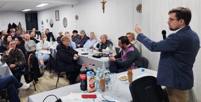 Mężczyzna w marynarce przemawia do zgromadzonych mieszkańców siedzących przy stołach, podczas spotkania w świetlicy wiejskiej.
