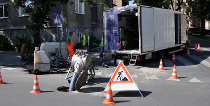 Ekipa pracowników w odzieży ochronnej przeprowadzająca prace związane z konserwacją kanalizacji na miejskiej ulicy, otoczoną pachołkami drogowymi i znakami ostrzegawczymi.