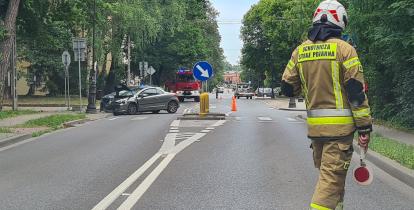 Strażak kieruje ruchem na miejscu wypadku drogowego, gdzie samochód osobowy uderzył w latarnię, a na miejscu obecne są służby ratunkowe.