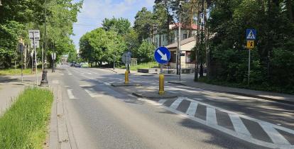 Ulica z wysepką rozdzielającą pasy ruchu, znakami drogowymi oraz przejściem dla pieszych, otoczona drzewami i budynkami.