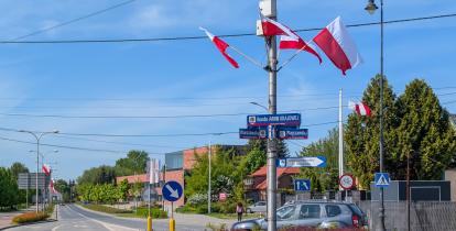 Rondo ozdobione polskimi flagami, otoczone znakami drogowymi i zielenią, znajduje się przy ulicy Warszawskiej w słoneczny dzień.