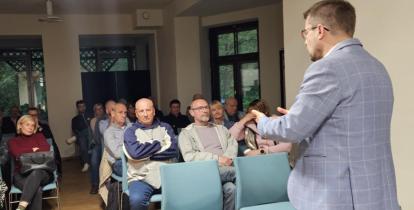 Mężczyzna w marynarce przemawia do zgromadzonych mieszkańców siedzących przy stołach, podczas spotkania w świetlicy wiejskiej.