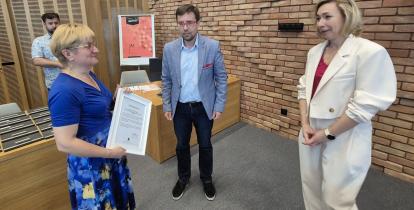 Kobieta w niebieskiej sukience odbiera podziękowanie w ramce od mężczyzny i kobiety podczas oficjalnego spotkania w urzędzie.