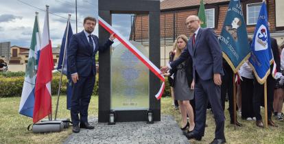 Uroczyste odsłonięcie pomnika upamiętniającego, z udziałem oficjeli i pocztów sztandarowych, odbywa się przy asyście flag narodowych i lokalnych.