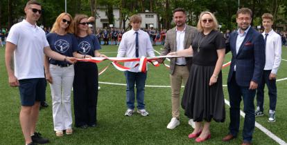 Grupa osób, w tym uczniowie i dorośli, przecina biało-czerwoną wstęgę podczas uroczystego otwarcia boiska sportowego.