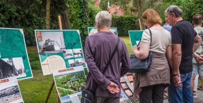 Grupa osób ogląda plansze z archiwalnymi zdjęciami i mapami podczas plenerowej wystawy historycznej. Ekspozycja ustawiona jest na sztalugach w parku, w otoczeniu zieleni. W tle widoczny ceglany budynek z czerwonym dachem.
